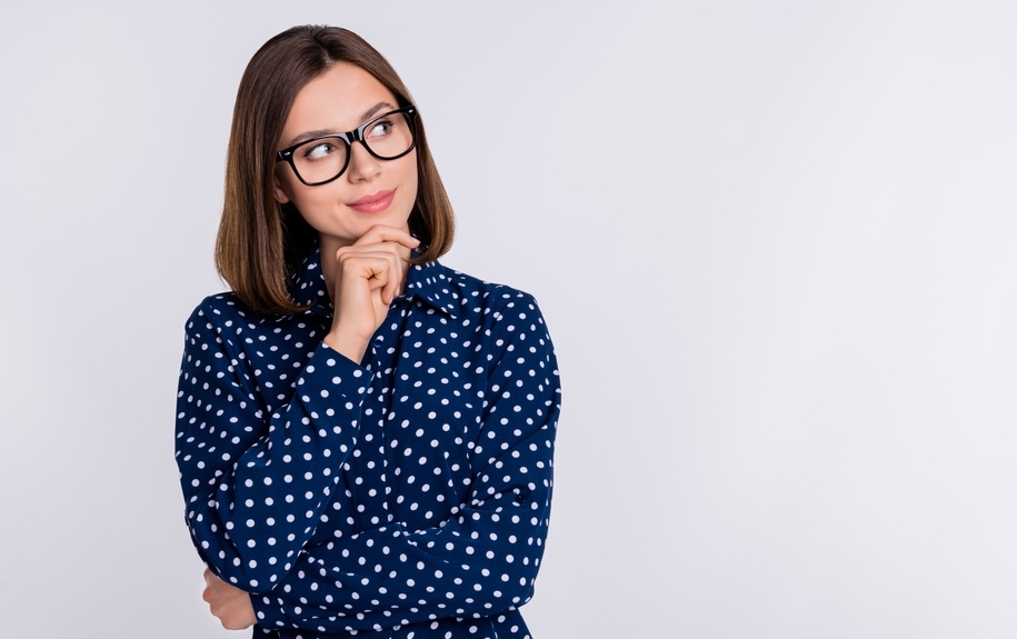 Thoughtful adult woman with neck-length hair and thick eyeglasses in a polka-dot shirt is depicted with her hand to her chin, looking upwards as if contemplating something. This image is being used in a blog titled "Am I a Candidate for Refractive or Laser Eye Surgery?" to illustrate the process of considering whether to pursue these types of surgeries.