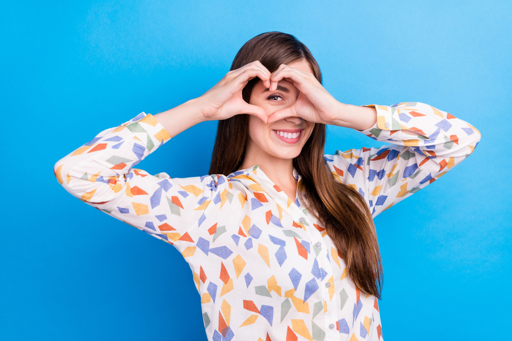 A woman with a bright smile is holding a heart-shaped gesture with her hands up to her face. 