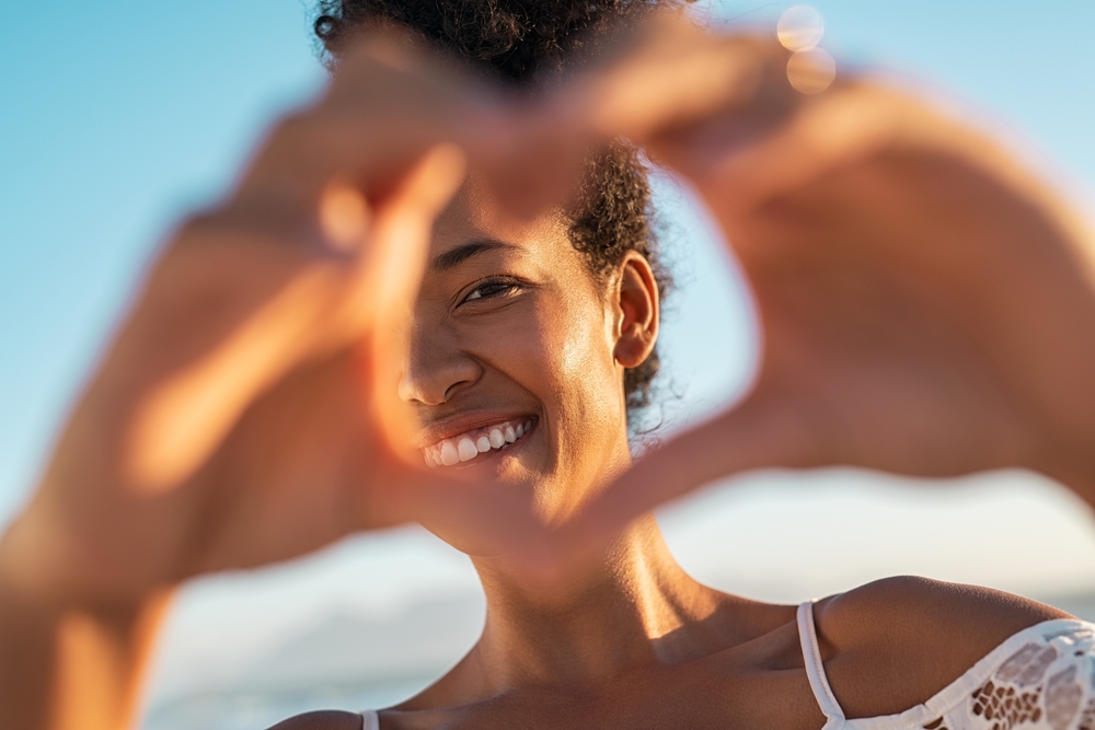 Young woman smiling and happy out in the summer time with her hands covering part of her face with a heart shape made with her hands.