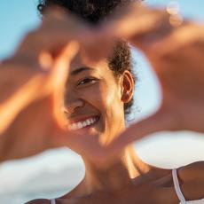Young woman smiling and happy out in the summer time with her hands covering part of her face with a heart shape made with her hands.